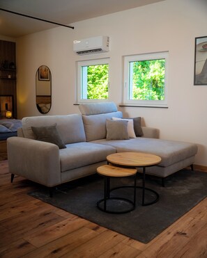 Ein modernes Wohnzimmer mit einem grauen Ecksofa, zwei runden Beistelltischen aus Holz, einem großen Spiegel an der Wand und zwei Fenstern mit Blick auf Grünflächen.