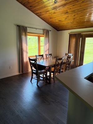 A dining area with a wooden table and chairs, a window with curtains, and a view of a grassy field.