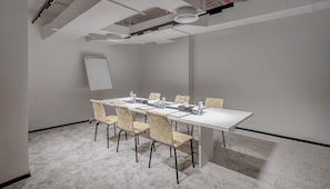 Uma sala de conferências com uma mesa branca, cadeiras bege, um quadro branco e piso coberto de carpete.
