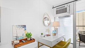 Habitación de hotel moderna con un escritorio blanco, una silla verde, un cuadro, una planta en maceta, una lámpara y una ventana con persianas.
