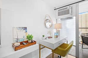 Een moderne hotelkamer met een wit bureau, een groene stoel, een schilderij, een potplant, een lamp en een raam met jaloezieën.