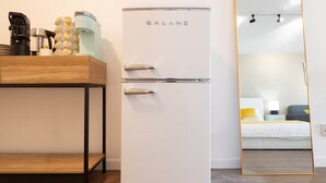Un refrigerador Galanz blanco en una habitación de hotel, con una mesa de madera y un espejo que refleja la habitación.