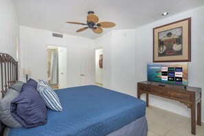 Een slaapkamer met een bed, een plafondventilator, een televisie op een houten standaard en een deur naar een andere kamer.