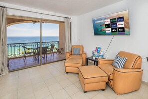 Een woonkamer met balkon en uitzicht op zee, ingericht met leren banken en fauteuils, een flatscreen-tv aan de wand met diverse streamingdiensten, en een bijzettafeltje met een decoratief object.