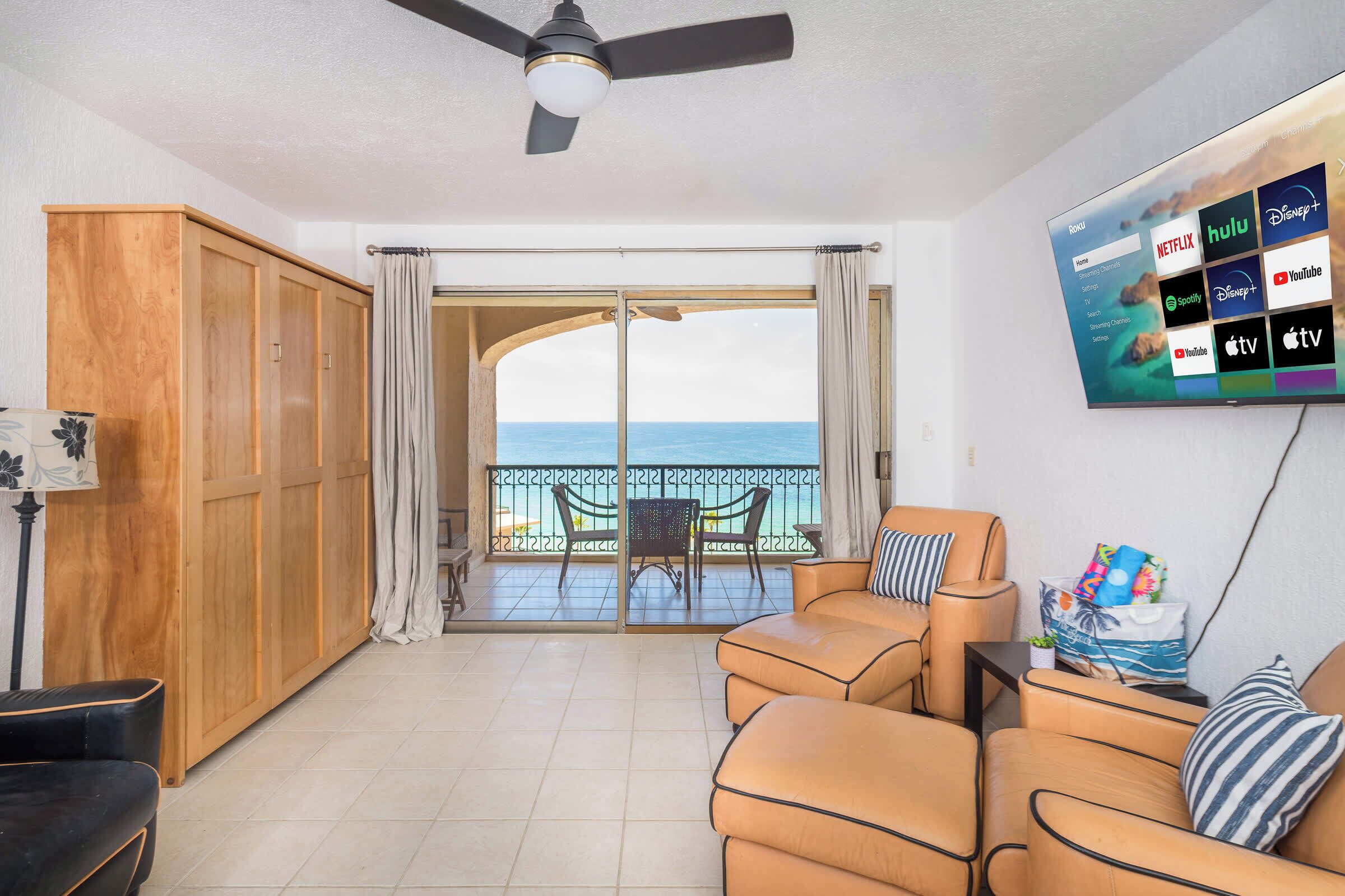 A living room with a sofa, a TV mounted on the wall displaying streaming services, and a balcony with a view of the sea.