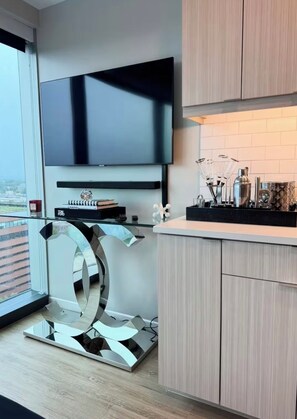 A modern kitchen with a flat-screen TV mounted above a countertop, a decorative mirror, and a view of the city through a large window. - Cozy 1-bedroom apartment in fantastic Raleigh with AC and gym access (Raleigh)