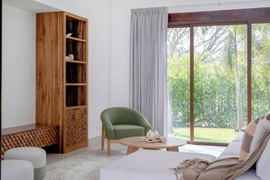 Ein modernes Zimmer mit einem grünen Sessel, einem weißen Sofa, einem Holzregal und einem großen Fenster mit Blick auf Grünflächen.