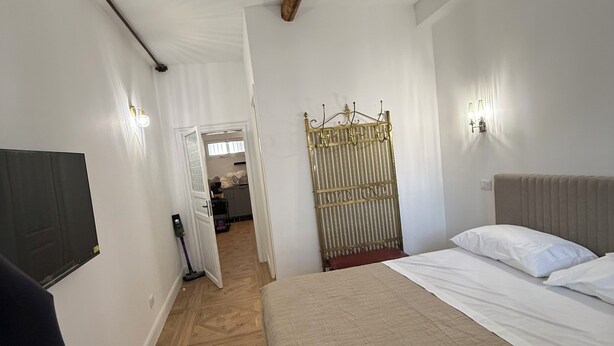 Quarto de hotel com cama, televisão fixada na parede, piso de madeira e porta que dá para um corredor.