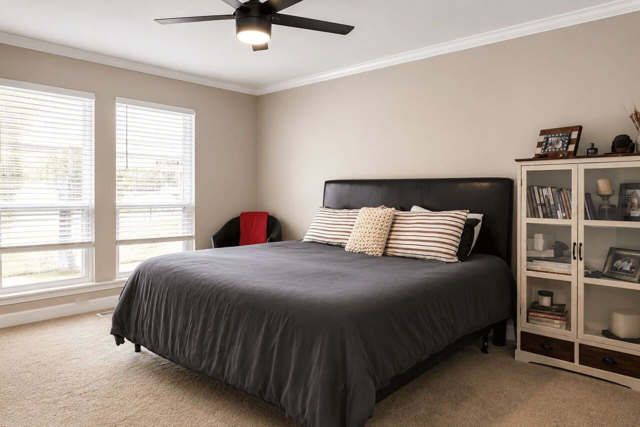 Quarto com uma cama, um ventilador de teto, uma janela com persianas e um armário com livros e objetos decorativos.