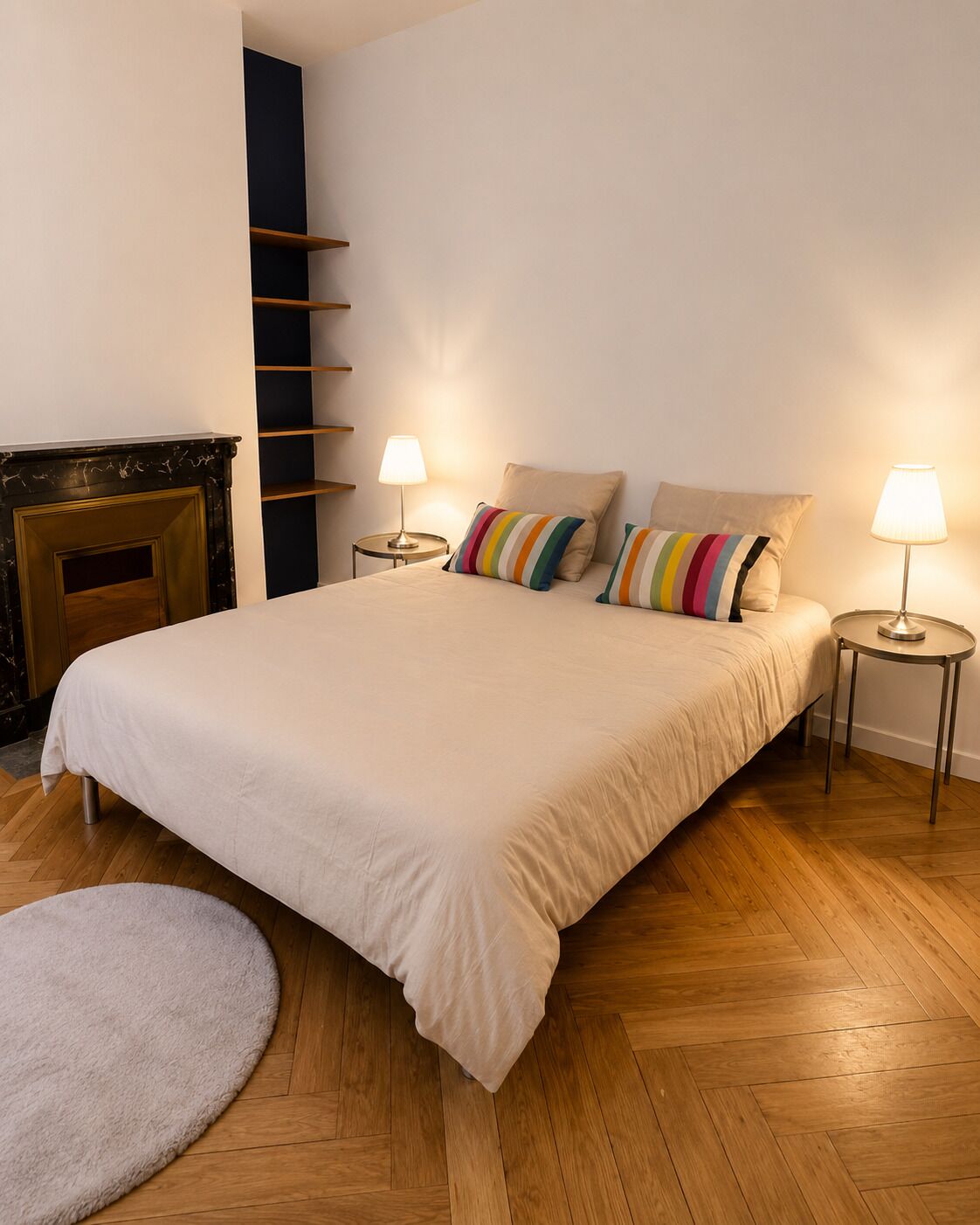 Une chambre à coucher avec un grand lit, deux tables de chevet avec des lampes, une cheminée et un parquet en bois disposé en motif à chevrons.