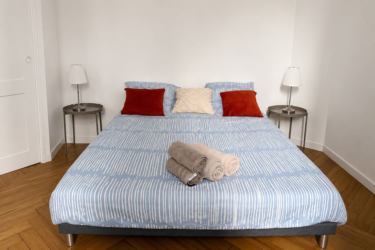 A neatly made bed with a striped bedspread, two red pillows, and a rolled towel on the bed. There are two bedside tables with lamps on either side of the bed.
