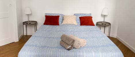 A neatly made bed with a striped bedspread, two red pillows, and a rolled towel on the bed. There are two bedside tables with lamps on either side of the bed.