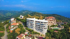 A coastal resort with multiple buildings, including a prominent white structure with balconies, surrounded by lush greenery and a view of the sea.