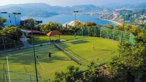 Uma quadra de tênis com vista para uma cidade costeira e montanhas ao fundo.