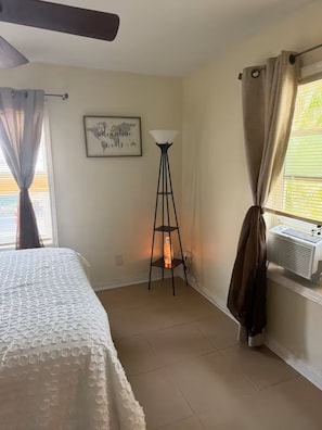 A bedroom with a bed, a standing lamp, a window with curtains, a wall-mounted air conditioner, and a framed quote on the wall.