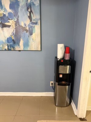 Ein Wasserspender mit Taschentuchbox und roten Bechern auf einer schwarzen Basis, ein blaues abstraktes Gemälde an der Wand und ein beige gefliester Boden.