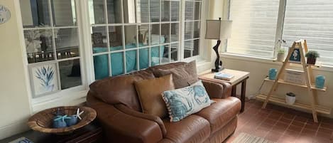 A living room with a brown leather sofa, a side table with a lamp, a wooden ladder shelf with decorative items, and a large window with blinds.