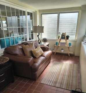 A living room with a brown leather sofa, a small table with a lamp, a fan, and a window with blinds.
