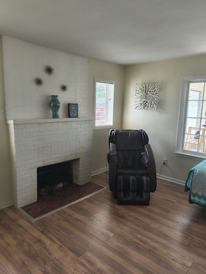 A room with a white brick fireplace, a massage chair, and a window with blinds.