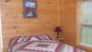 A wooden cabin room with a bed, a picture on the wall, and a window with blinds. - American Pines Cabin #5 (Keystone)