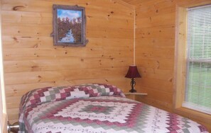 A wooden cabin room with a bed, a picture on the wall, and a window with blinds. - American Pines Cabin #5 (Keystone)