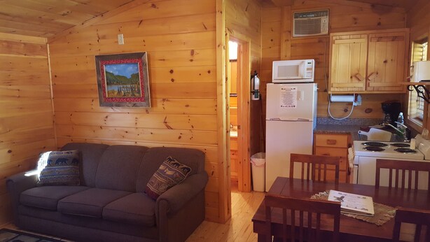 A cozy cabin interior with a wooden interior, a sofa, a dining table, and a kitchen area including a refrigerator and microwave. - American Pines Cabin #5 (Keystone)