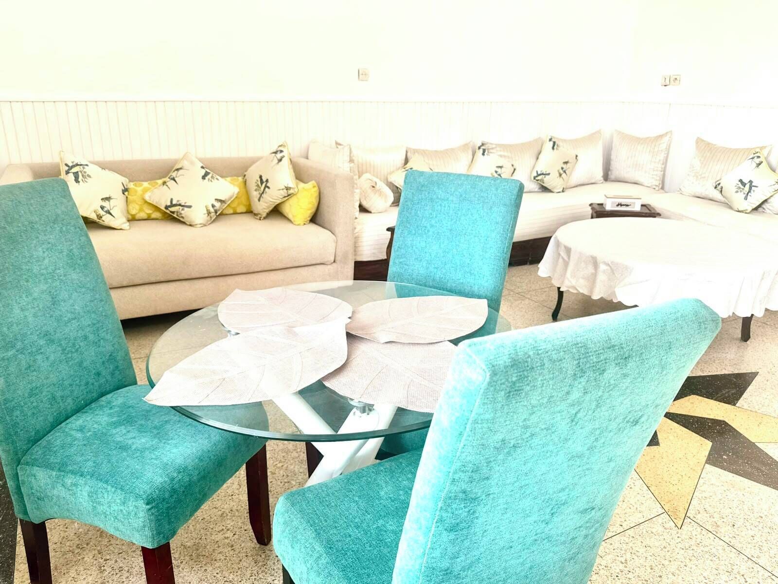 A room with a round glass table, teal chairs, a beige sofa with patterned cushions, and a white bed with a white tablecloth.