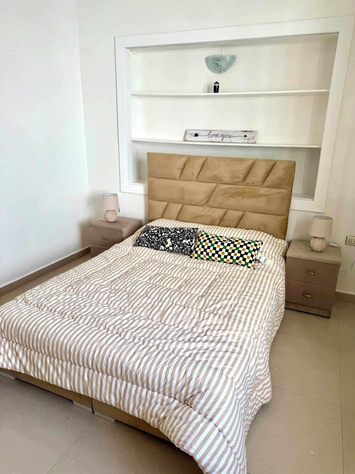 Een slaapkamer met een bed, nachtkastje, ladekast, wandplank met decoratieve objecten, en een wandklok.