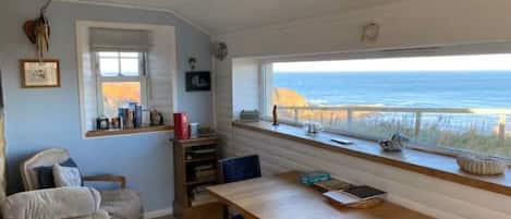 Una habitación con vista al océano, una mesa de madera, un sillón con cojines y una estantería.