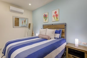 Une chambre à coucher comprenant un lit, une table de chevet, une commode, un climatiseur et deux tableaux accrochés au mur.