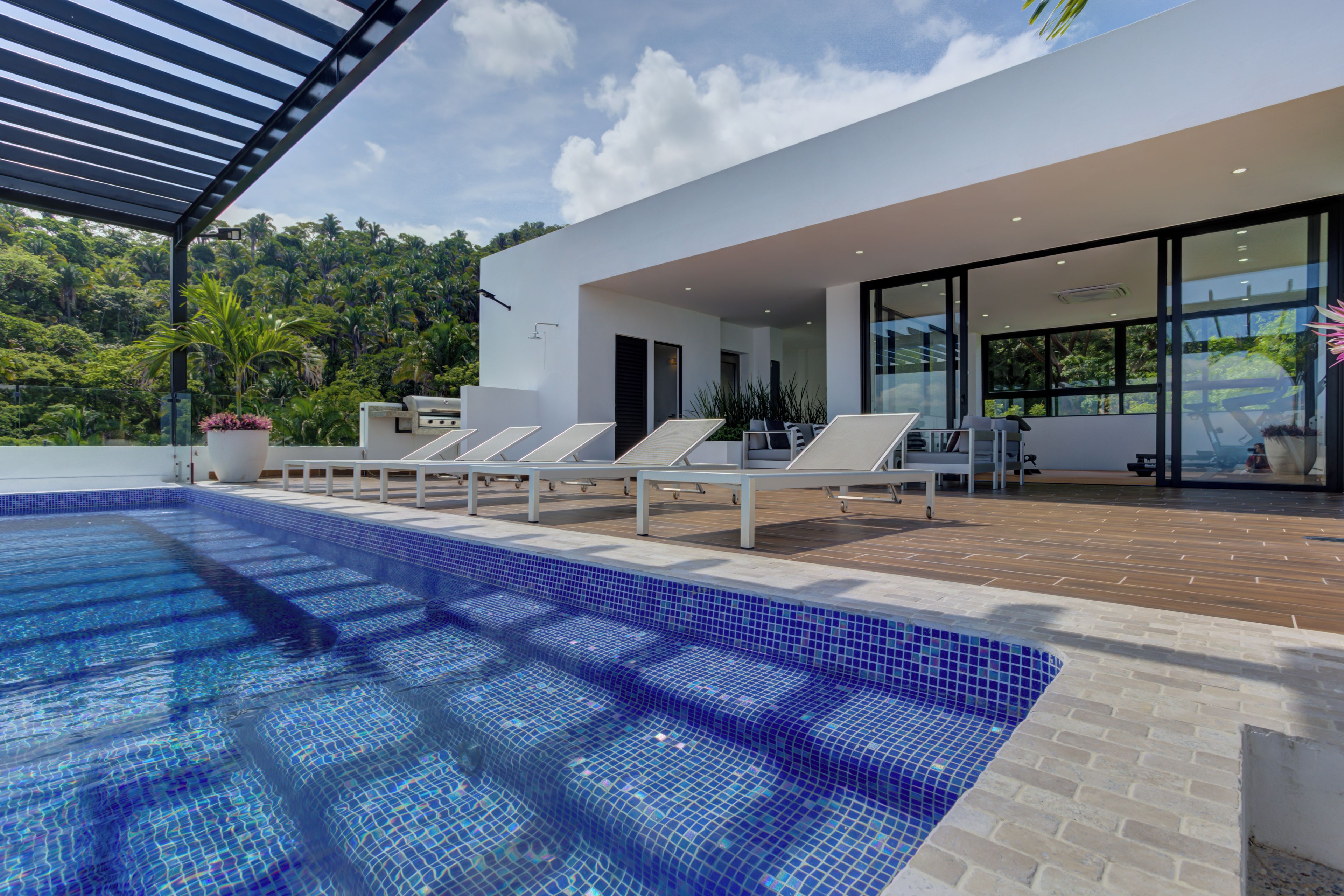 A modern outdoor pool area with lounge chairs, a wooden deck, and a view of lush greenery.