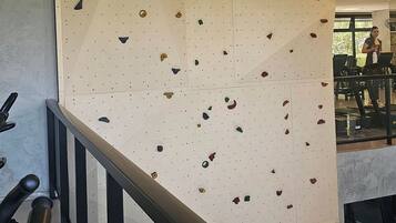 Uma parede de escalada com vários pontos de apoio, um corrimão e uma pessoa utilizando uma esteira ao fundo.