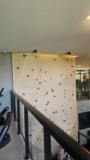 Uma parede de escalada com vários pontos de apoio, um corrimão e uma pessoa utilizando uma esteira ao fundo.