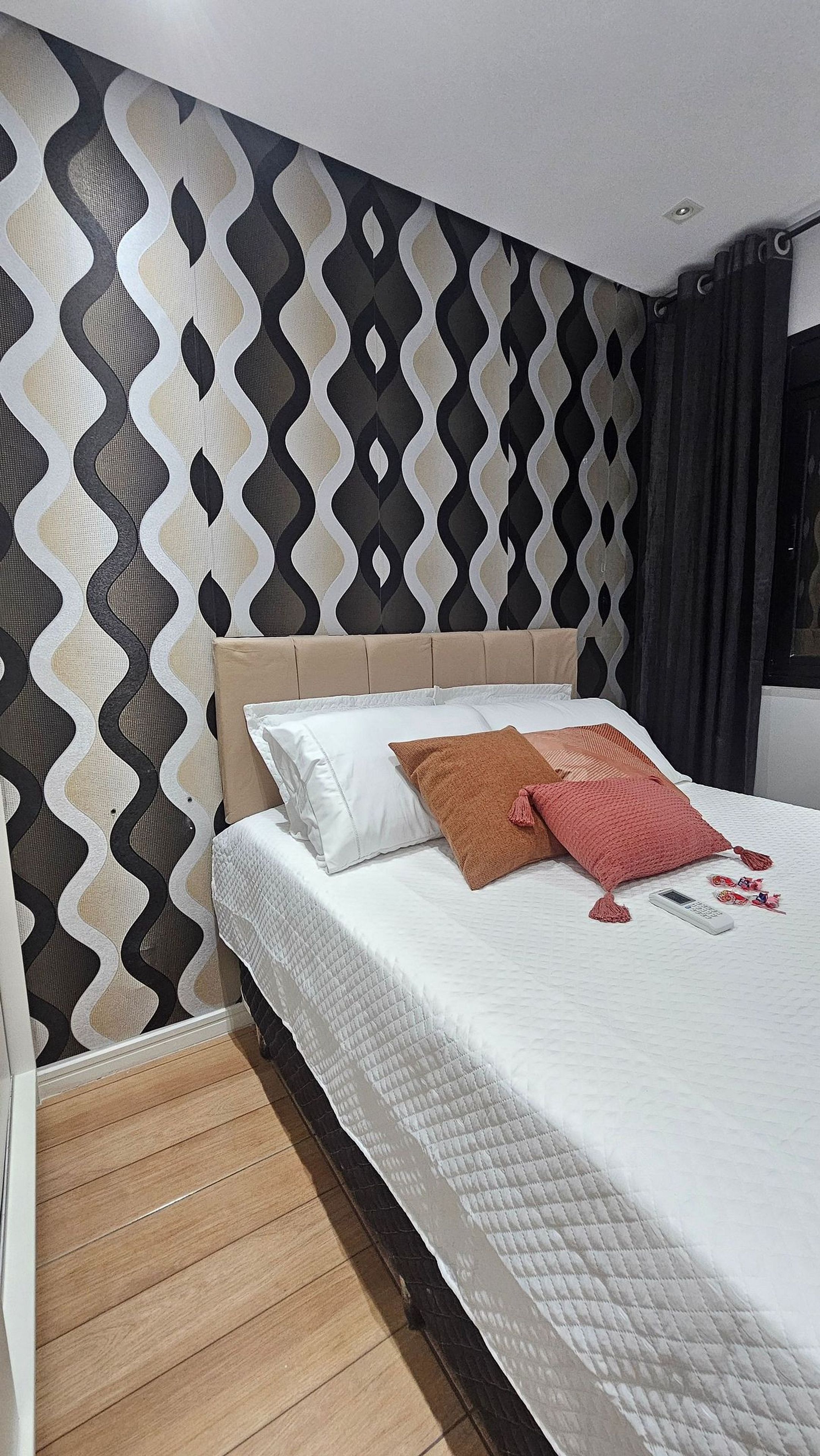 Quarto de hotel com parede estampada, cama com roupa de cama branca e chão de madeira.