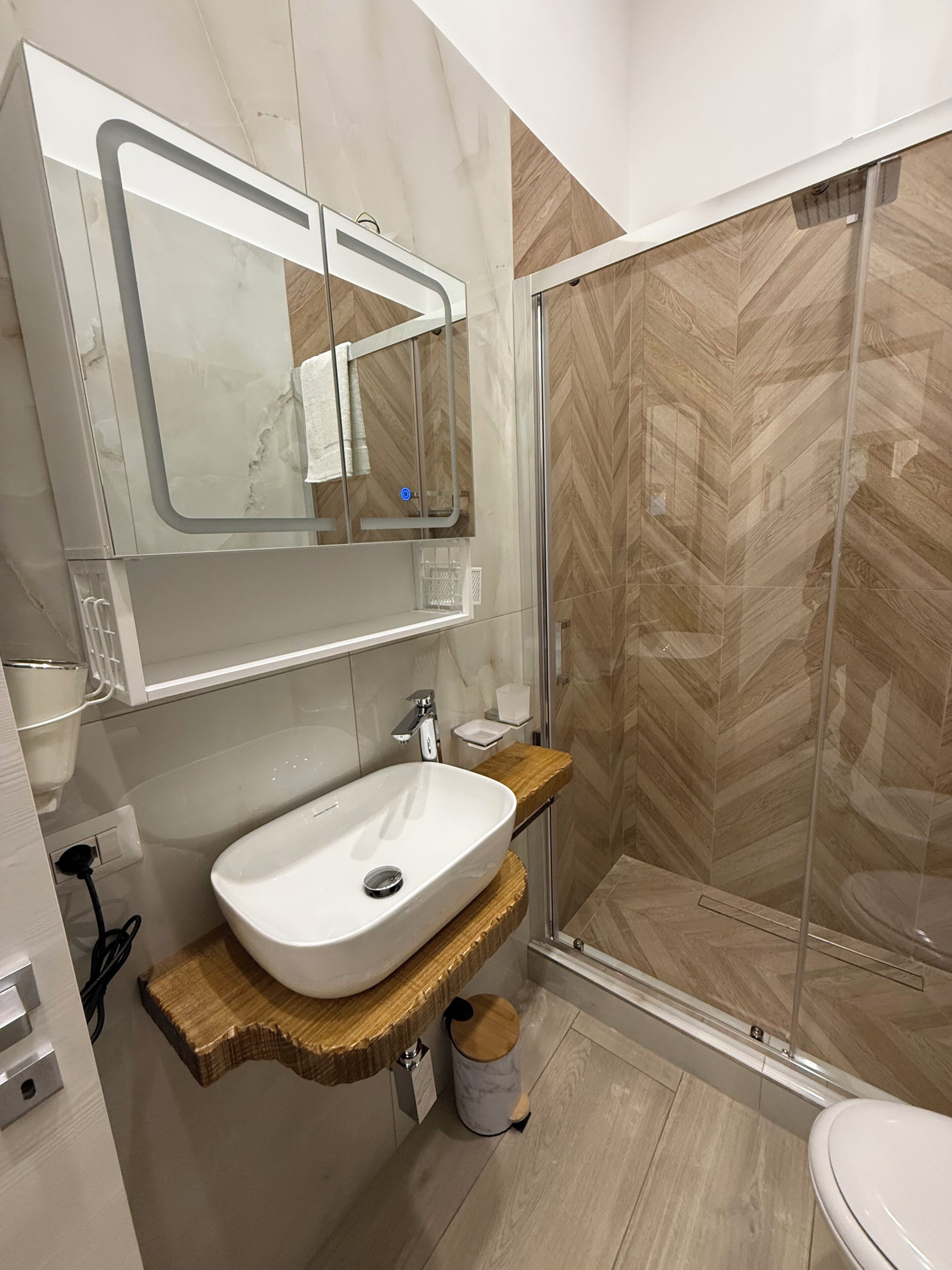 Une salle de bain avec un lavabo blanc, un meuble-lavabo en bois, un miroir et un espace douche dont le sol est recouvert d’un motif en chevrons.