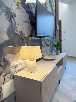 Ein modernes Wohnzimmer mit Marmorwand, einem flachbildfernseher über einer beigen Ablage, einer Lampe und einer Zimmerpflanze.