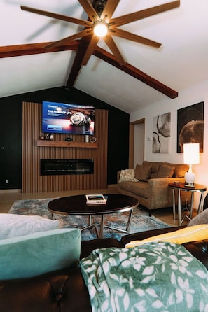 A living room with a television mounted on a wooden wall, a ceiling fan, a sofa, a coffee table, and a lamp. - Airport Family Getaway Pvt. Pickleball/Game Room (San Antonio)