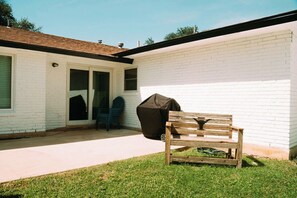Ein Garten mit einer Holzbank, einem überdachten Grill und einem weiß verputzten Gebäude mit Tür und Fenstern.