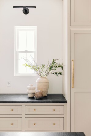 Una cocina con un armario blanco, una encimera negra, un florero con flores y una ventana.