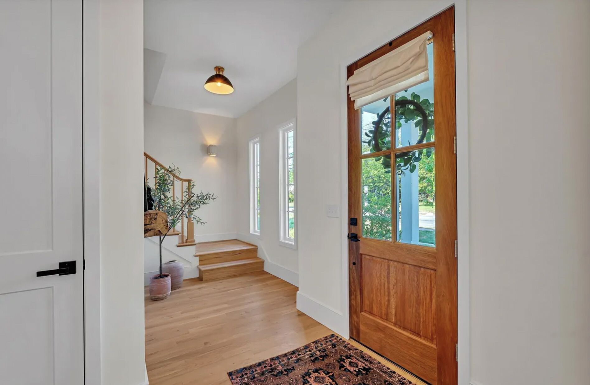 A hallway with a wooden door, a staircase, and a view of greenery outside.