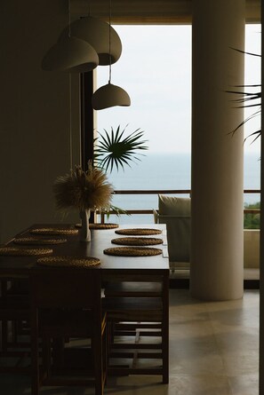 A dining area with a view of the ocean, featuring a table with woven placemats, hanging pendant lights, and a vase with dried plants.