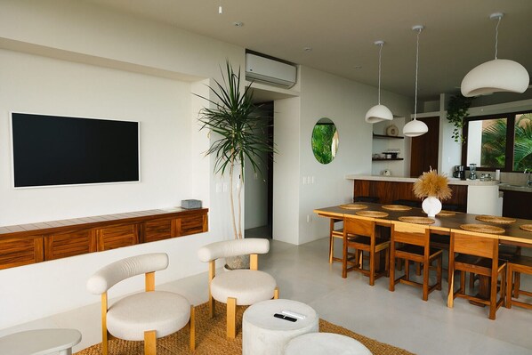 A modern living room with a flat-screen TV, a dining area with a wooden table and chairs, a large potted plant, and a kitchen with pendant lighting.
