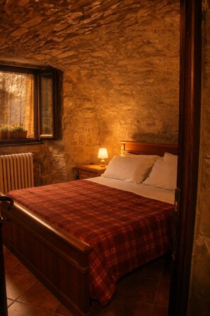 A bedroom with a stone wall, a wooden bed frame, a red plaid bedspread, a bedside lamp, and a window with curtains. - A sustainable and eco-friendly stone house near the Cinque Terre. (Villecchia)