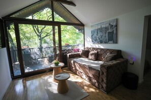 Una habitación con un ventanal grande, un sofá marrón, una mesita redonda con una planta y un cuadro de paisaje urbano en la pared.