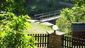 Un muro de piedra con una verja metálica, un pequeño puente sobre un canal de agua y abundante vegetación.