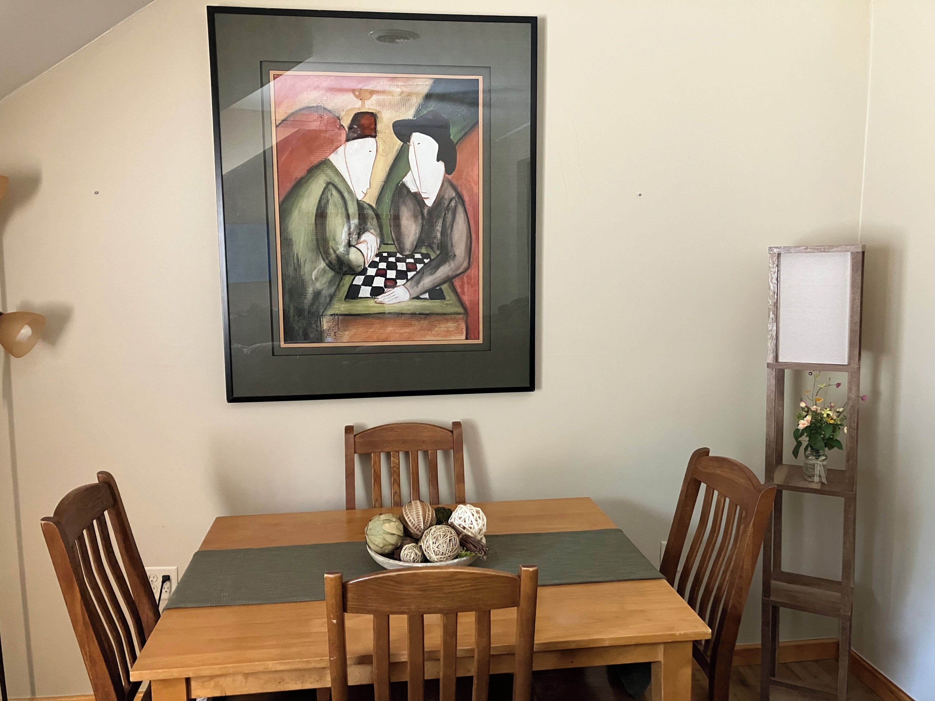 A dining area with a wooden table, four chairs, a framed artwork, and a small vase with flowers on a side table.