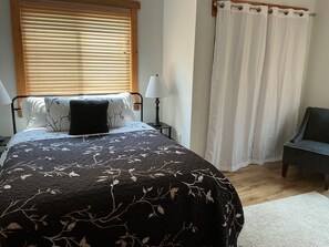 A bedroom with a bed, two bedside lamps, a chair, a window with blinds, and a curtain.