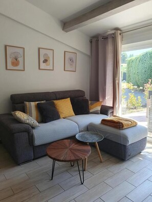 Ein modernes Wohnzimmer mit einem grauen Sofa, einem kleinen runden Tisch und Blick auf einen Garten durch ein großes Fenster.
