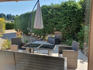 Een terras buiten met rieten meubels, een tafel met glazen blad en een hangende parasol.