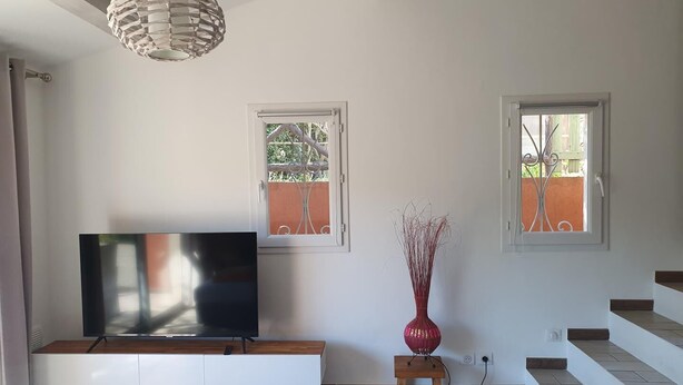 A living room with a flat-screen TV on a white cabinet, a red vase on a small wooden stool, and a staircase to the right. - La Belle Aupsoise - AUPS (AUPS)